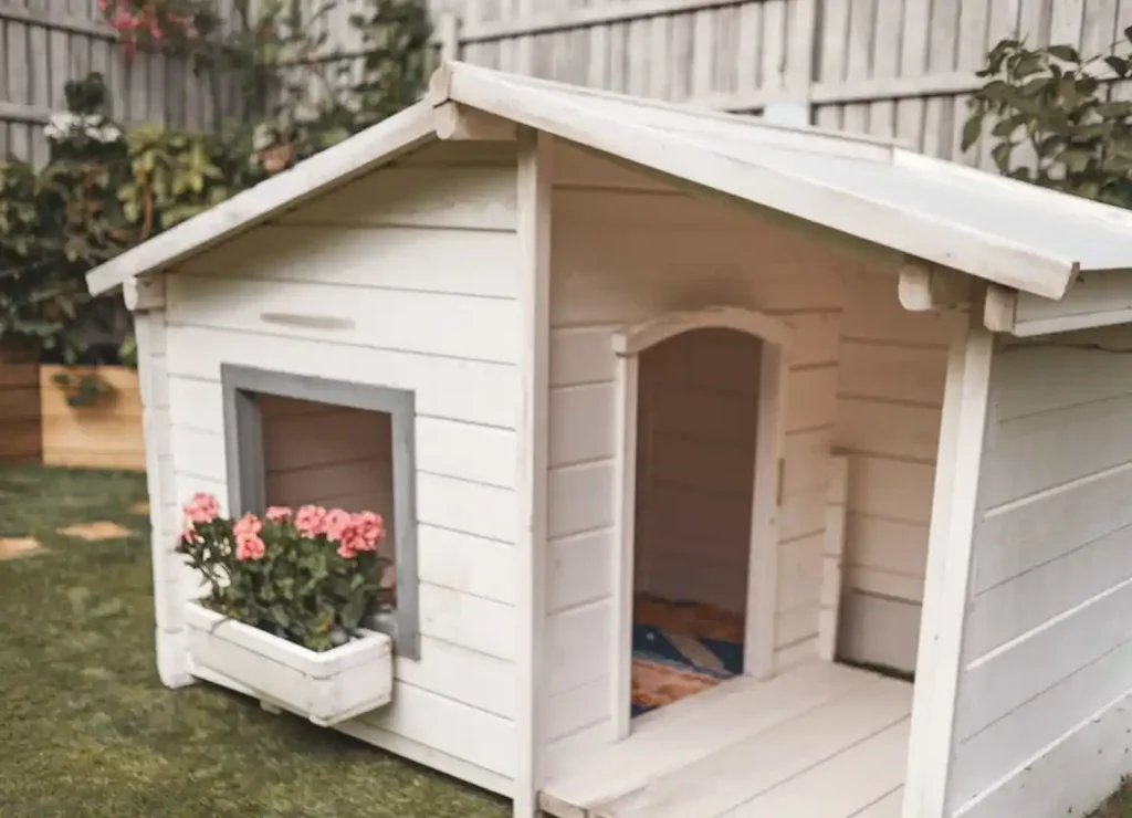 White cottage-style dog kennel with pink flowers in window box and covered porch